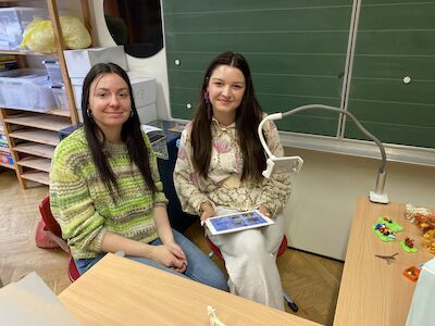 Zwei junge Frauen sitzen mit einem Tablet und buntem Bastelzeug an einem Schreibtisch.