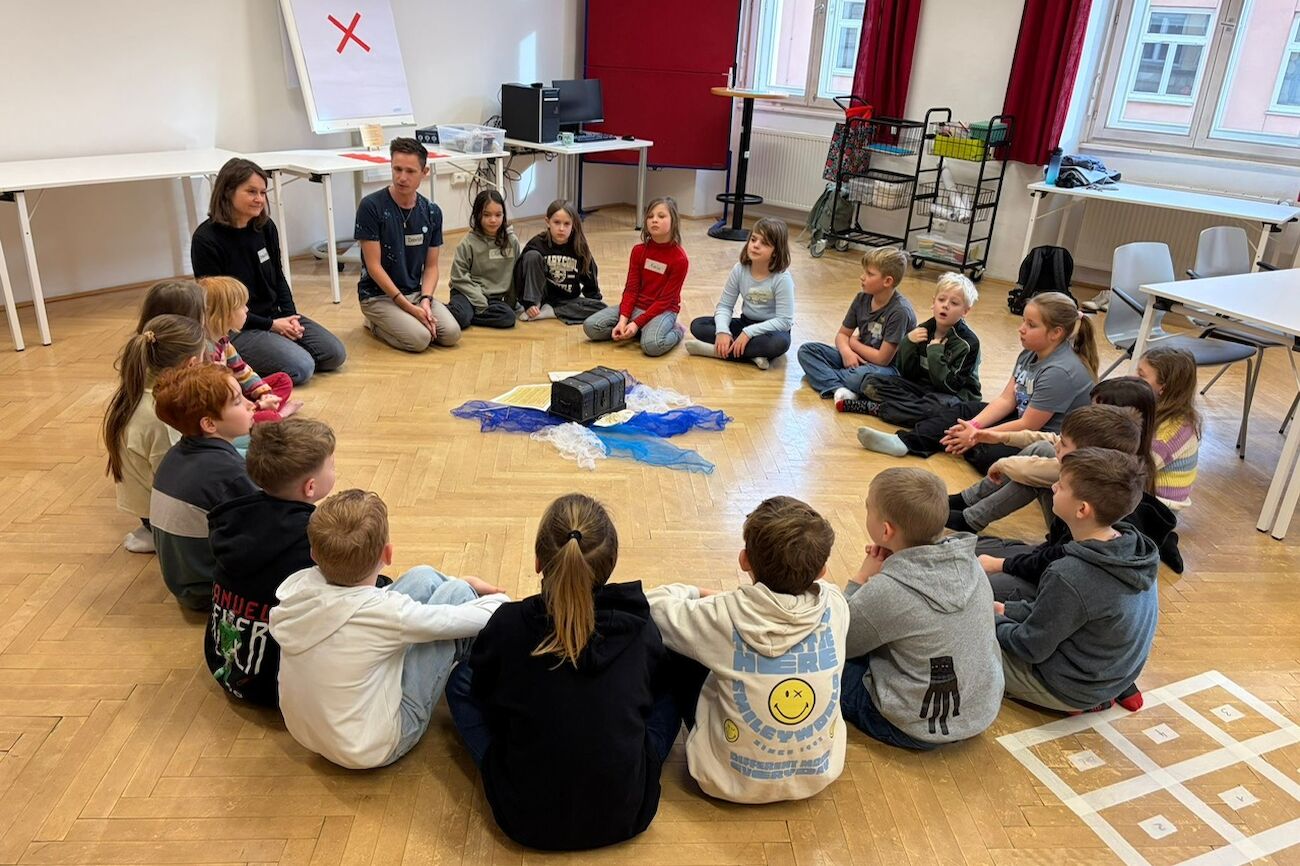 Kindergruppe mit Lehrenden sitzend am Boden des Seminarraumes
